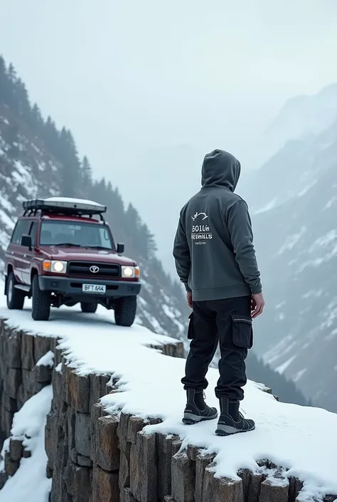 A man standing on a cliff  in winter facing toward a valley with a land cruizer beside him and the car is maroon and its plate number  is ,"BFT 684" and the man is wearing a hoodie with a writing written as,"5010~ReDHILLs.COM" and he is wearing a boot with...