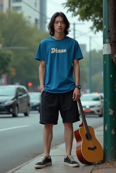 The image shows a handsome korean young man ,long hair,standing on a sidewalk next to a tree. He is wearing a blue t-shirt with word "Dimas" writted, black shorts, and black sneakers. He has a guitar leaning against a green pole next to him. The background...