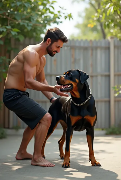 Wearing black shorts, interacting with a Rottweiler dog. He is outdoors, in a backyard with a concrete floor, surrounded by a wooden fence and a white wall in the background. The dog wears a chain collar and is looking at the mans hand, apparently interact...