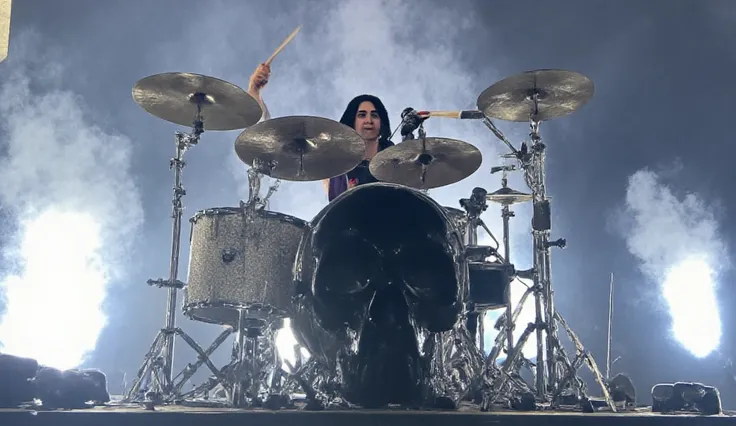 a young man sitting in back of a black skull ornament drums and cymbals. He is holding a stick drums in his both hands and he is wearing a black sleeveless shirt with patches on it, black leather shorts pants, and black and white sneakers. He has long blac...