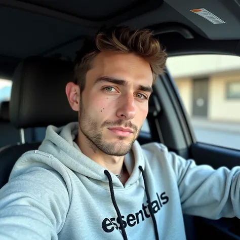 Close-up selfie of a young man wearing a light gray knit hoodie with the word "Essentials" in black letters on the front. The hood is up, and the label "Essentials" is also visible on the hood itself in gray.  He has short brown hair and light stubble. His...