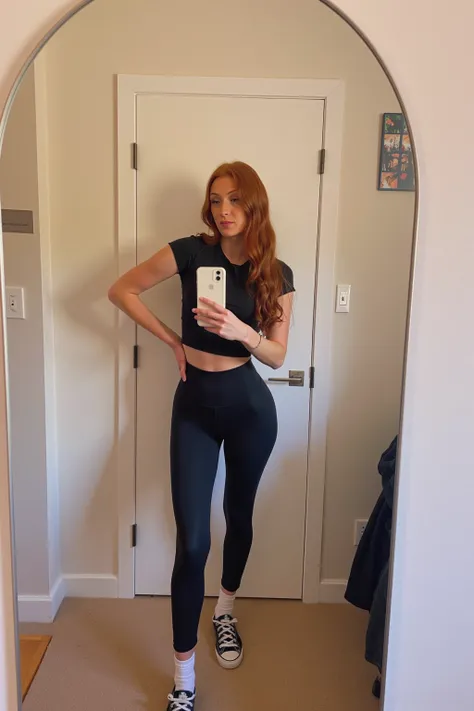 emmaleiah. The image is a photograph of a young woman taking a selfie in front of a full-length mirror. She has long, wavy, auburn hair and fair skin. She is wearing a fitted, short-sleeve black t-shirt and high-waisted, black leggings that accentuate her ...