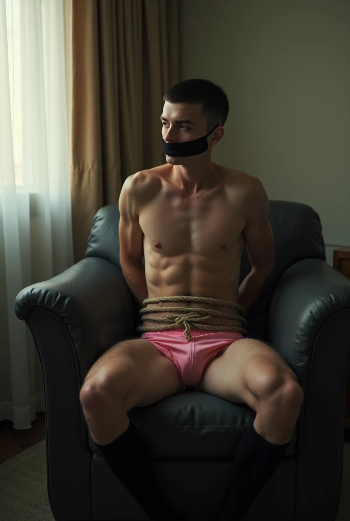 A 20 year old Russian guy with dark buzz cut hair is sitting in a chair in the hotel room. He is wearing pink mini Tanga briefs and black socks. His hands are behind his back and his feet are tied together with rope. His mouth is covered with black tape. 