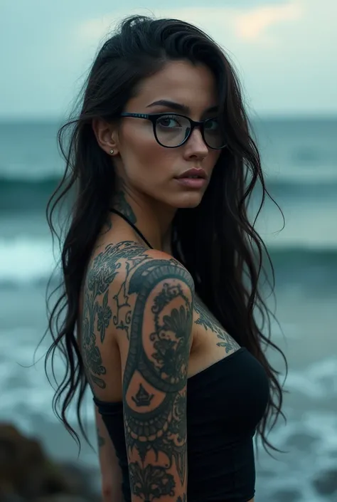 A tattooed woman with glasses, with long wavy hair on the back, in front of the sea