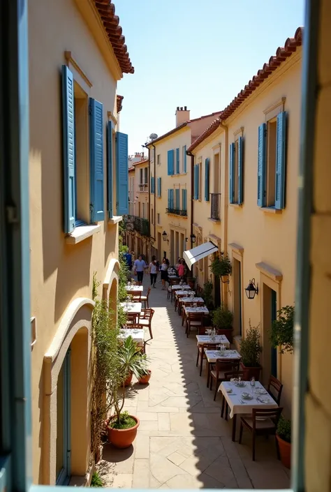  A view from the second floor of a house , from the balcony ,  over a Mediterranean-style street .  The perspective offers a wide view of the light beige buildings with blue shuttered windows,  lined up along the paved street .  The tables and chairs of an...