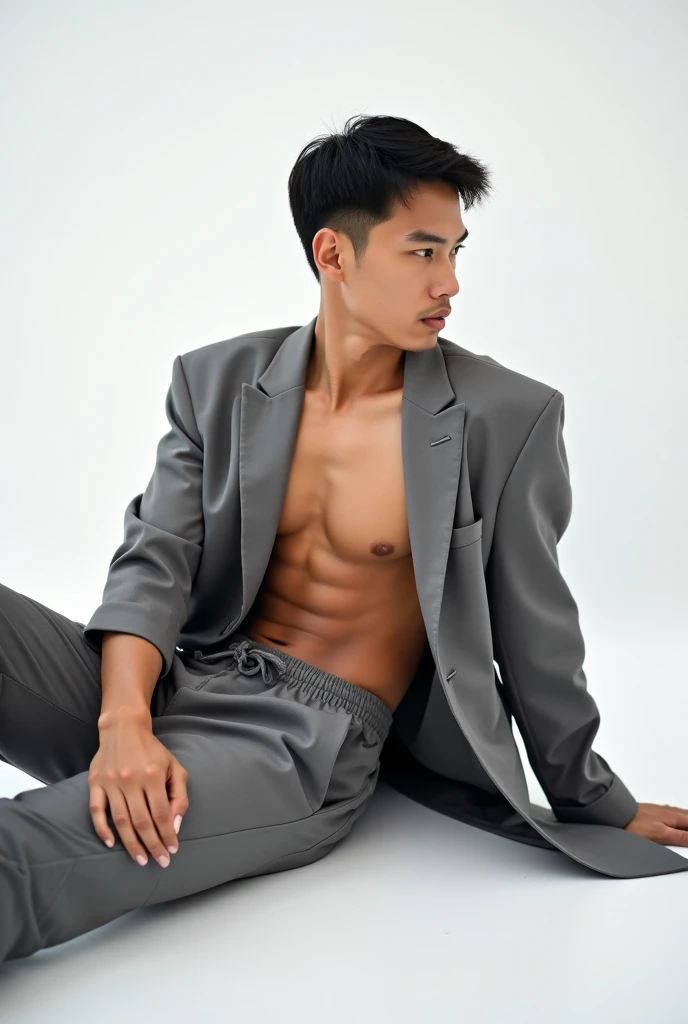 A young, muscular Asian man with short black hair and tan skin sits on a white studio floor. Hes wearing an open, oversized gray suit jacket and matching loose-fitting pants, which reveal his bare chest and the top of his gray waistband. His pose is relaxe...