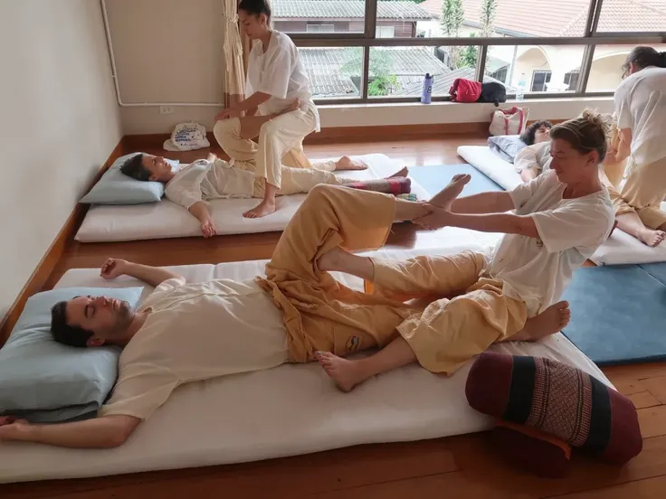 An indoor Thai yoga massage class with natural sunlight streaming through large windows. The room has wooden floors and mats arranged in rows, with students practicing Thai yoga massage in pairs. Each person is dressed in loose, traditional massage attire ...