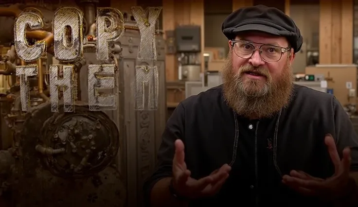 The image features a bearded man wearing a black hat, glasses, and a black shirt with stitched detailing. He is gesturing with his hands in a conversational manner. On the left side of the image is a large, vintage industrial machine with a circular metall...