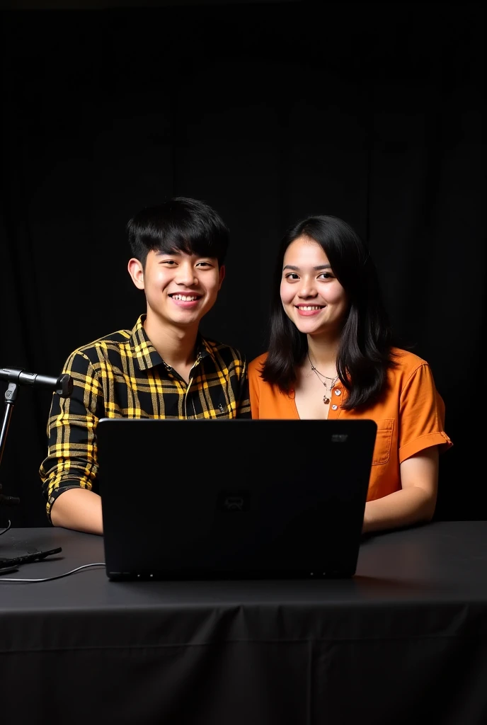 A 21-year-old boy sits in front of a computer desk with a microphone and a laptop. He wears a black and yellow checked shirt and has black hair against a black themed background. And sitting next to him is his girlfriend who is also 21 years old.