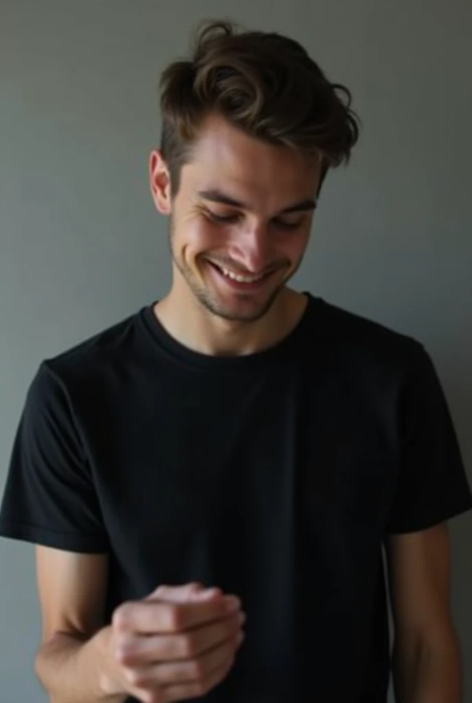 A close-up of the upper body and face of an attractive young man wearing a black blank t-shirt  smiling slightly while looking down at his hand that is holding something. The background is a gray wall, the lighting is soft, the colors are muted, the mood i...