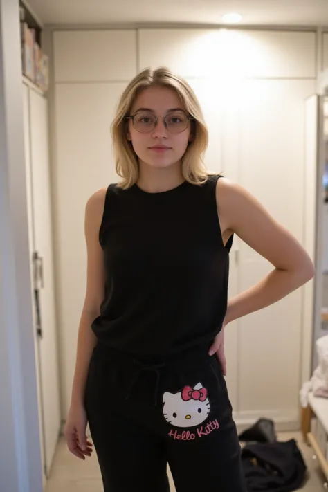 The photo shows a blonde woman . in glasses.She is wearing a black top and black Hello Kitty pants.  In the background you can see a white wardrobe and a door .  There is a dark jacket or sweater on the floor .  The room is lit by soft light .