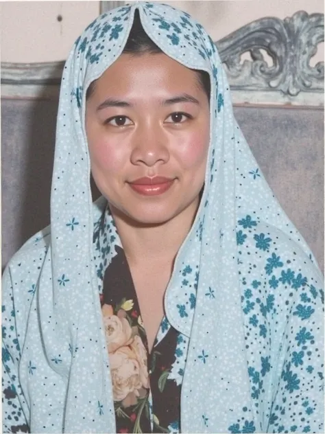 The image shows a person wearing a light-colored, batik-patterned hijab over a dark-colored blouse. The background features a wooden chair with floral decorative elements.