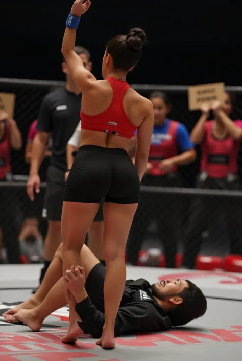 A victorious grappling tournament scene on a mat in a jiu-jitsu gymnasium. An Asian woman with a fit and athletic physique stands confidently, wearing a red racerback sports bra and black athletic shorts. She poses with her back slightly turned toward the ...