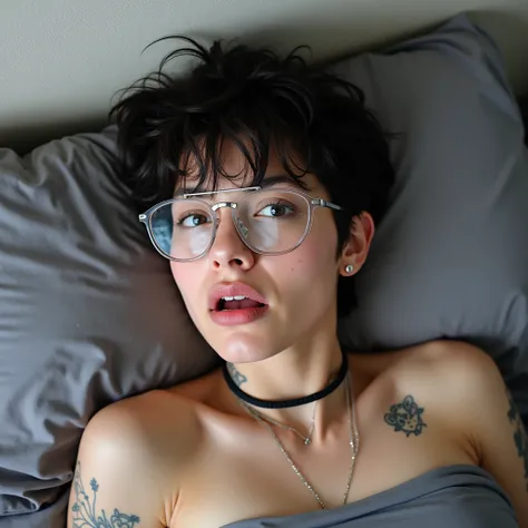 The image features an individual lying down on what appears to be a bed with gray covers. This person has several visible physical characteristics: short dark hair, a nose piercing, multiple tattoos on the arms and shoulders, and a choker-style necklace. T...