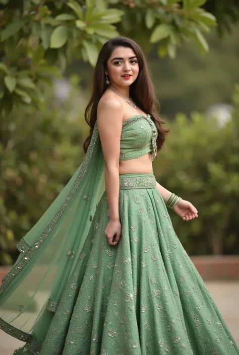 A female model poses gracefully outdoors, wearing a traditional green lehenga with intricate patterns and a matching crop top. Her long hair cascades down, and she is adorned with delicate jewelry, including a nose ring and a pendant. The background is fil...