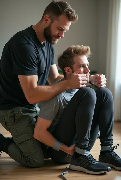 A nordic man with trimmed beard and short hair dressing cargo pants, t-shirt and black long riding boots has tied up a blond man wearing Adidas running shoes who is sitting on a floor hands behind back.  The nordic man has wrapped gray tape tightly around ...