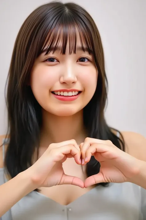 A close-up shot of a woman standing and making a heart shape with her hands. She has a warm and gentle smile, with her eyes looking directly at the viewer. Her hands are positioned in front of her chest, forming a perfect heart. The background is soft and ...