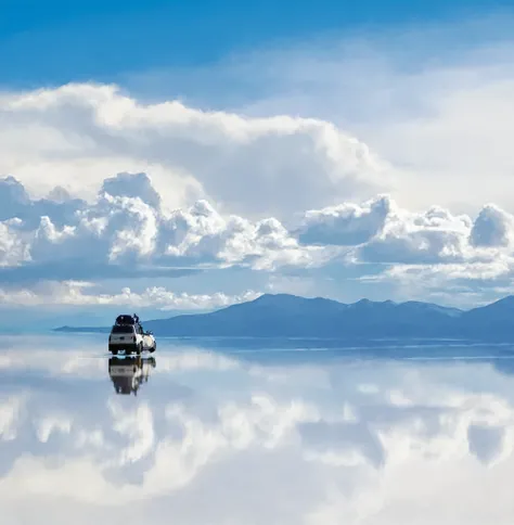 A white SUV with roof racks glides across the still snow mountain, its reflection merging with the mirror-like surface. The deep blue sky glows with the northern lights, their green and purple hues dancing above. Wisps of clouds and distant gray-blue mount...