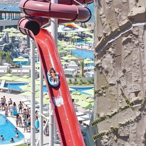 "A high-angle, diagonal view of a massive red water slide, appearing broken and dangerous. The slide has large cracks and pieces falling off mid-air, making it look unstable. Yellow CAUTION maintenance tapes are placed along the descent, warning of danger....