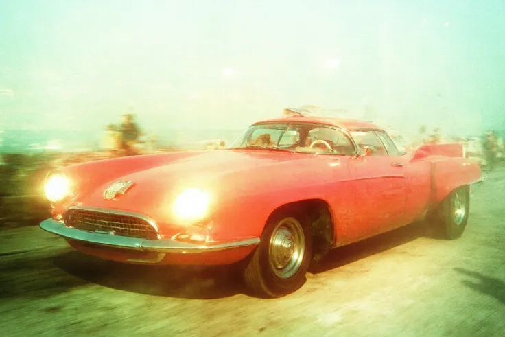 A red car with headlights on driving down the road, a photo of an art deco-style 1950s American sports car, a blurry shot in the style of Roger Fretaille of people in a vintage Miami Beach club, a light green and pink color palette, a foggy and misty atmos...