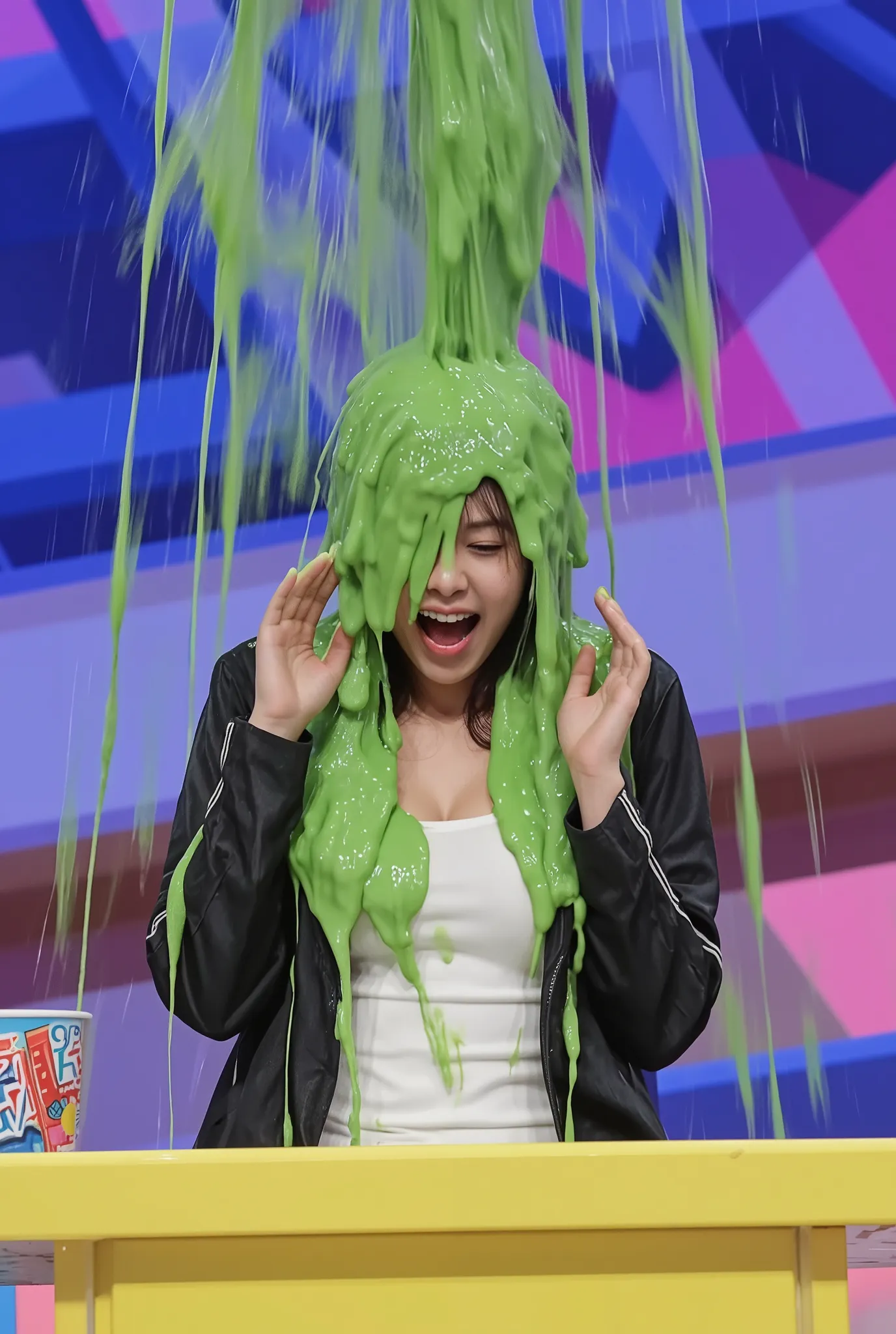 A lively and colorful moment captures a Korean woman seated on a stage, getting drenched in a thick cascade of vibrant green slime pouring from above. The individual, wearing a white shirt with cleavage and a black jacket, reacts with raised hands and an e...