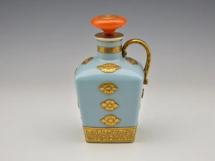 A spirit bottle. Square bottle, rounded corners. Pale blue glass. bright orange inlaid geometric swirls at top. Gold rim, gold aztec inspired pattern near bottom. Gold labels. Gold lid. There is a handle on the neck 