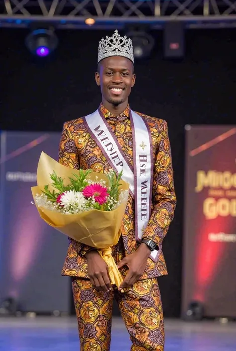 A young male beauty pageant winner standing on stage, wearing a stylish African print outfit with elegant details. He has a confident smile, a sparkling crown on his head, and a sash across his chest that reads ‘Mister Gbeke’. He holds a bouquet of flowers...