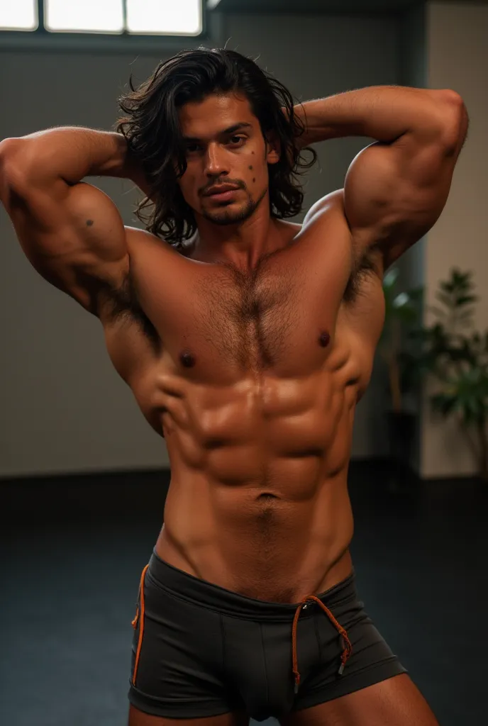 Latin Mexican man tanned and brown hairy 17-year-old muscular athletic man with his arms behind his head, velvety armpits, wearing shorts and bulgo in the gym