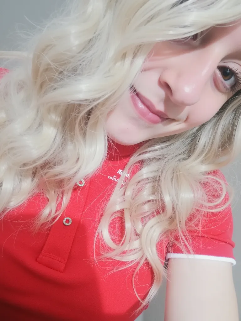 Close-up portrait of a cute, playful girl with long, wavy blonde hair and fair skin. She has brown eyes and a soft, cheerful smile. She's wearing a red polo shirt with a 'YouTube eyacuteangel' text on it. Her face is slightly tilted towards the camera, sho...