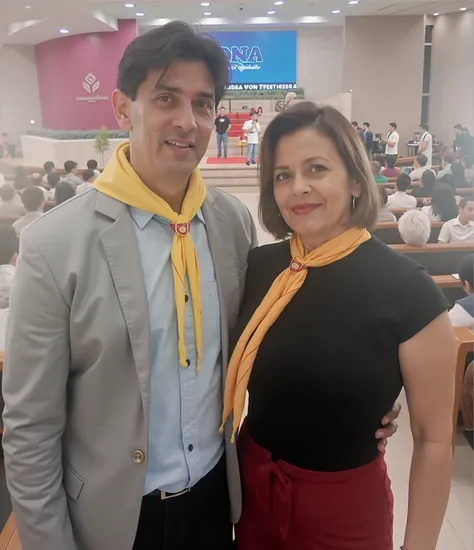 A man and a woman stand side by side in a bright, modern interior. The man, of Hispanic descent and in his 30s, wears a light gray blazer over a blue patterned shirt, accessorized with an all-yellow Boy Scout-style scarf. He has short, dark hair and a conf...