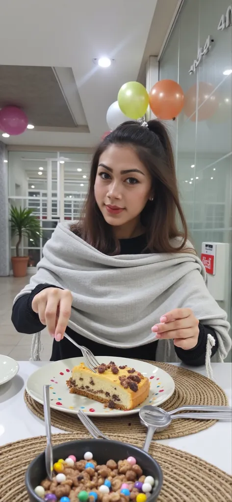 Young woman sitting at a table with a plate of food in front of her. she is wearing a gray shawl over her shoulders and has a fork in her hand. the plate has a slice of cake on it and there is a bowl of cereal on the table next to it. the woman is smiling ...