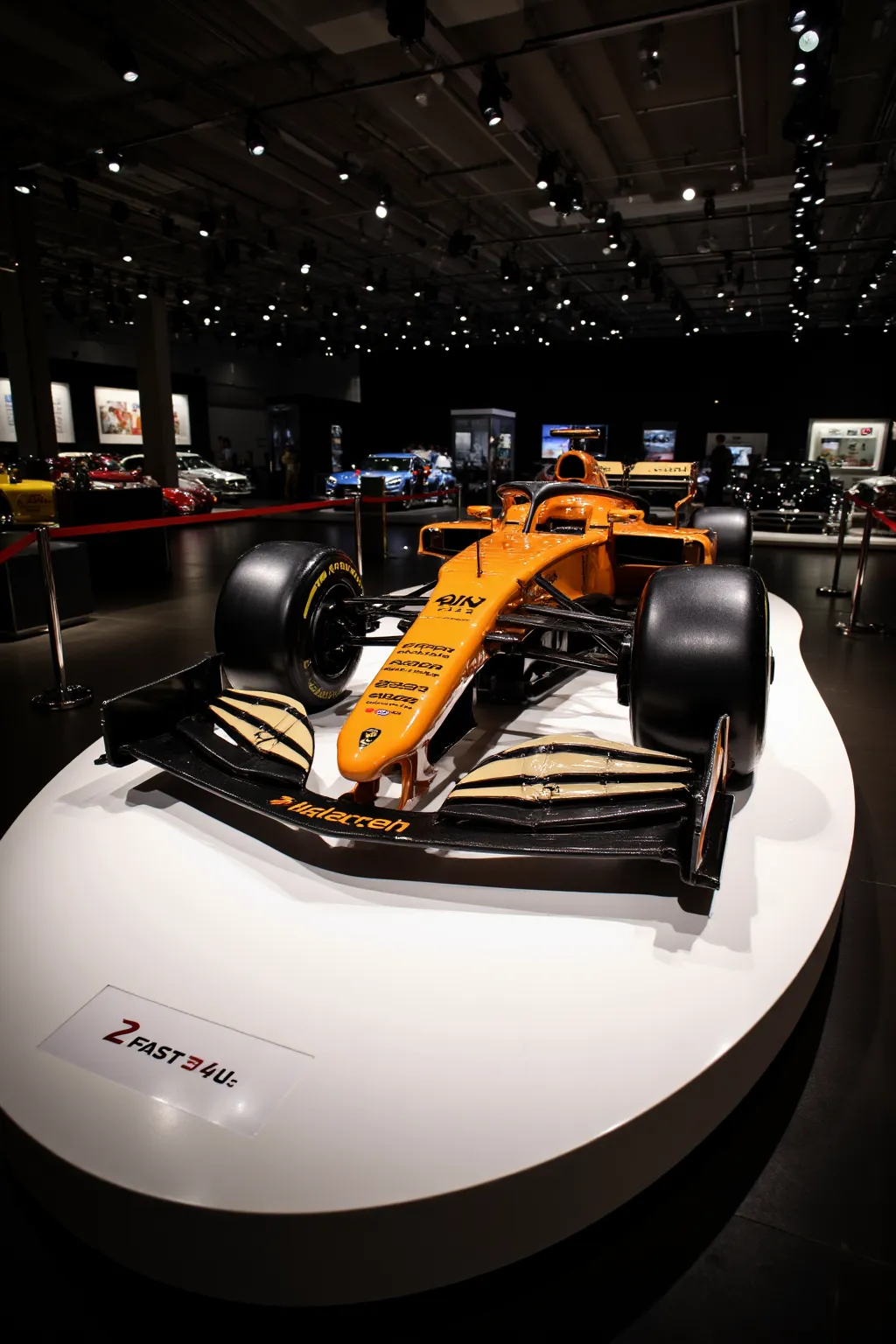 Shiny McLaren Formula 1 car. BREAK In a Showroom with spotlights focusing on the car. There is a velvet rope around the parameter of the car. Car is on a white, ovular disk in the center of the show room. There is a sign on the ovular disk that reads "2FAS...