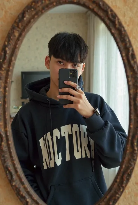 A young man of approximately  with black hair, Between collar and smooth, takes a selfie in front of a mirror with his phone. He wears a dark hoodie with the word "victory" printed in large, visible letters. The image is color. The mirror has an ornate fra...