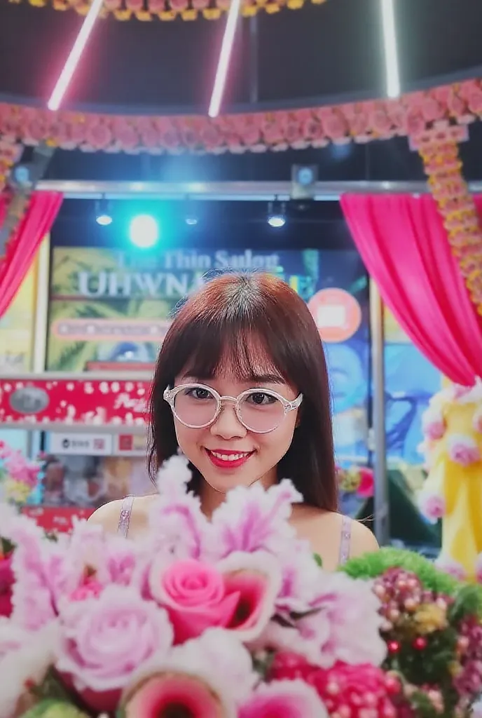 The image of a brunette, a 25-year-old Thai woman standing on stage in a pink dress with a white Westground overlay. She has long bangs that look smooth and healthy. She wears round silver-rimmed glasses that complement her stylish look and wears a Casio B...