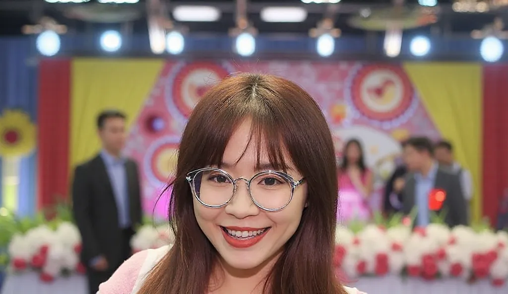 The image of a brunette, a 25-year-old Thai woman standing on stage in a pink dress with a white Westground overlay. She has long bangs that look smooth and healthy. She wears round silver-rimmed glasses that complement her stylish look and wears a Casio B...