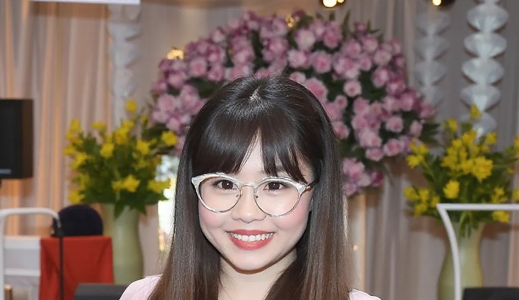 The image of a 25-year-old Thai woman standing on stage in a pink dress with a white Westground overlay. She had long, slick bangs that looked soft and healthy. She wore round silver-rimmed glasses that complemented her stylish look and wore a white Casio ...