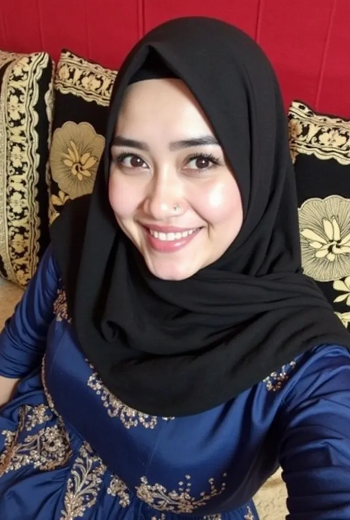 The image features a woman wearing a black hijab and a blue dress adorned with golden embroidery. She appears to be taking a selfie with a gentle smile. Her facial features are clear, and her eyebrows are drawn in a dark shade. The background includes cush...