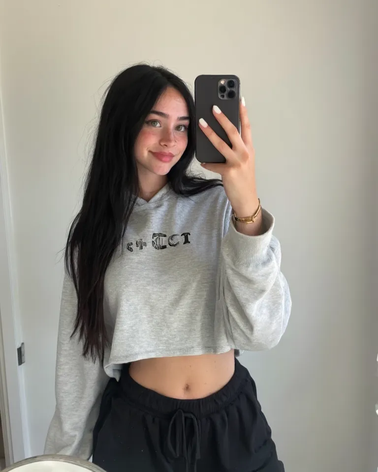  S3lfi3 1girl, solo, green eyes and black very long hair, long shiny hair, dressed in a stylish crop top hoodie, wearing black sweatpants, photo beside a wall suggests a clean, in a room minimalistic aesthetic that allows her features and outfit to stand o...