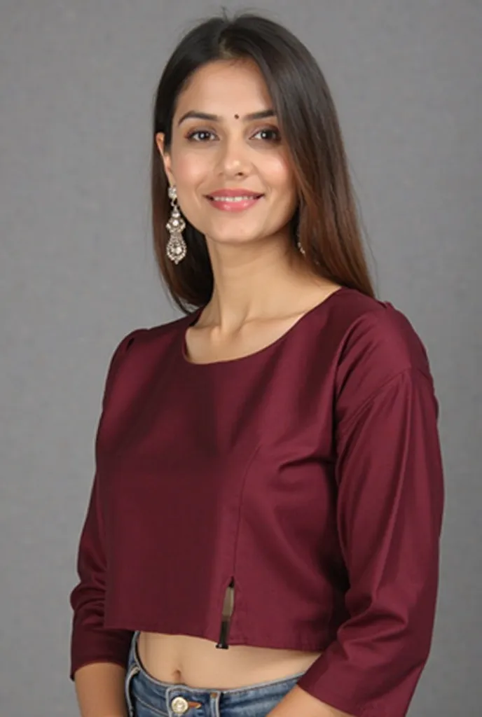 "A young woman with a confident smile poses in a stylish maroon saree blouse against a neutral gray background. She has long, straight brown hair neatly tucked behind her ears, revealing elegant drop earrings. The blouse features a modern round neckline, t...