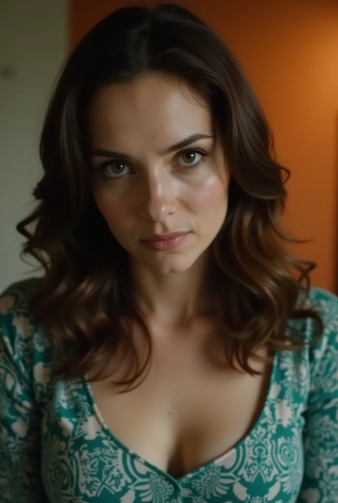 A close-up of a woman with shoulder-length brown hair, a teal and white patterned top, and a slightly downturned expression, captured in low-light indoor settings with a slightly orange-toned wall behind her, focusing on the subject's face and chest area.
