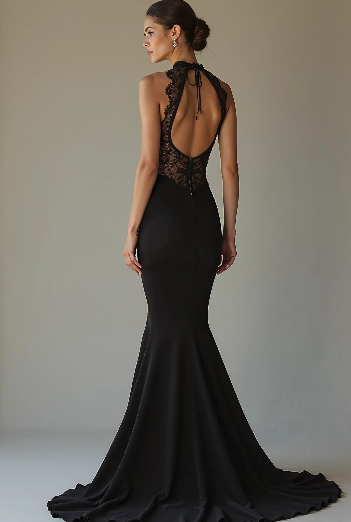 A slender model radiates sophistication in a stunning black evening gown, featuring an elegant lace detail that stretches from around her neck down to the gown. The dress boasts a backless halter design, with delicate straps tied at the neck, accentuating ...