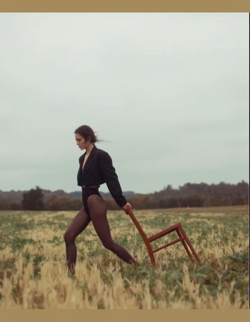 create an image identical to this one just change the hair to long A cinematic fashion editorial shot of a woman walking through an open field, elegantly dragging a wooden chair behind her. She wears an oversized black blazer, slightly open, revealing a bl...