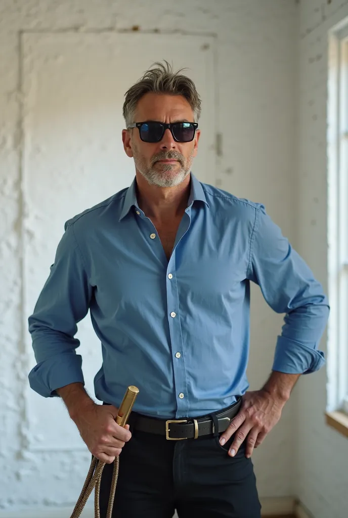 A handsome man in his early forties stands in a room with white walls, wearing a blue shirt , sun glasses and black pants. His left hand is on his hip while his right  hand holds a whip The man's expression is focused and  serious