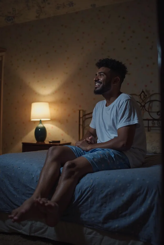 Profile photo of a young man wearing blue sleeping shorts and white short sleeve t-shirt sitting on his bed with his feet on the floor looking happy and illuminated by the light of a lamp on the nightstand in Venezuela, Guarico state, on a dark night. 