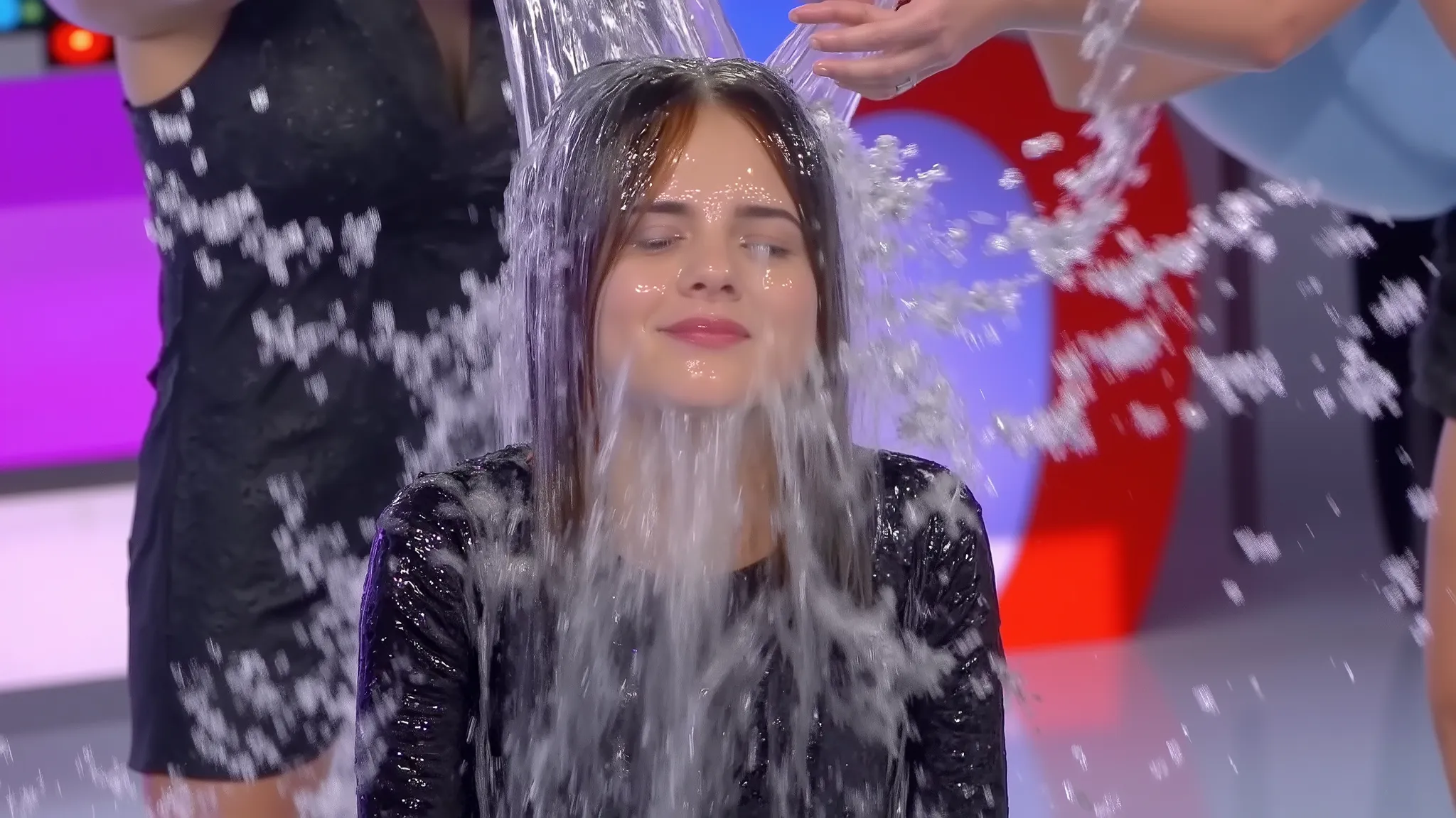 On the set of a TV gameshow that is elaborately decorated with lots of set dressing and brightly lit with lots of coloured lights, a beautiful 18-year-old half black, half white mixed race girl wearing a sparkly black dress, jewellery, lipstick and makeup ...