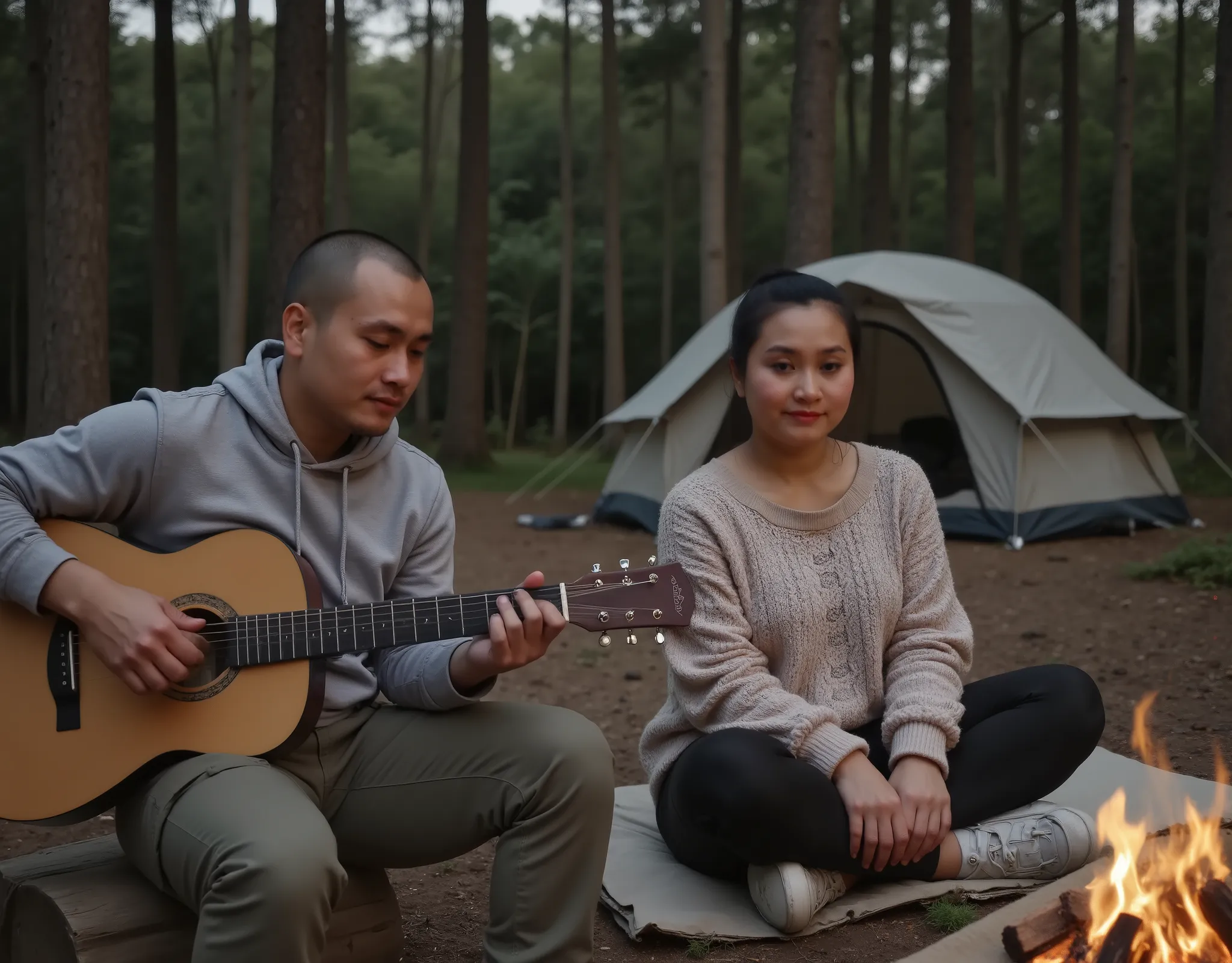In a quiet forest clearing in West Java, an Indonesian couple sits closely beside a small campfire under the darkening sky. The man, with a nearly bald head and a clean military-style haircut, wears a gray hoodie and cargo pants. He’s seated on a log, casu...