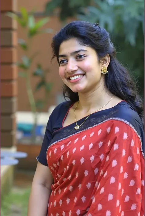 A woman is standing outdoors against a blurred background of green foliage and a brick wall. She is wearing a vibrant red sari with a pattern of white shapes, paired with a black blouse that has short sleeves. Her dark hair is styled in loose waves cascadi...