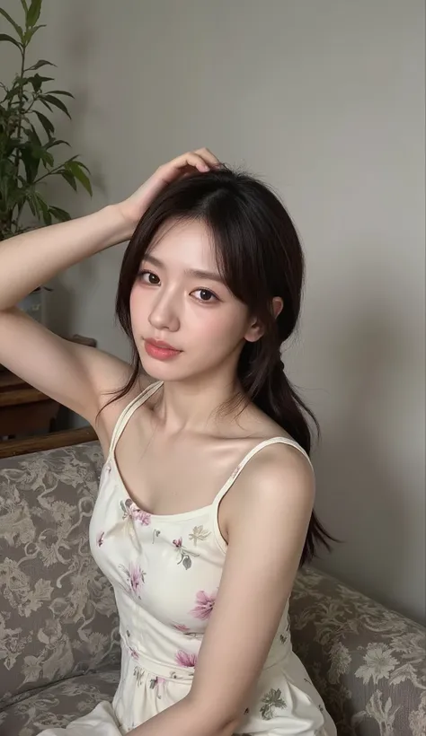 A young woman is sitting comfortably on a patterned couch, her posture relaxed and casual. She wears a light, floral-patterned sleeveless dress, which adds a touch of elegance to her appearance. Her left arm is raised, as she gently holds her dark hair bac...