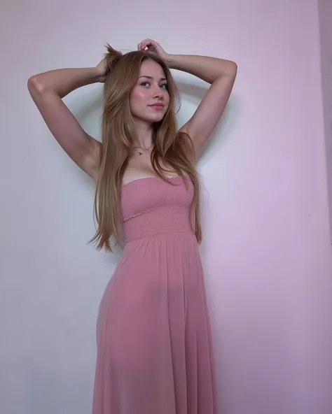 A young woman with brown hair and a soft expression strikes a pose wearing a pink dress, her arms raised behind her head, against a neutral backdrop. The photograph captures a single figure against a minimalist backdrop, the subject positioned slightly off...