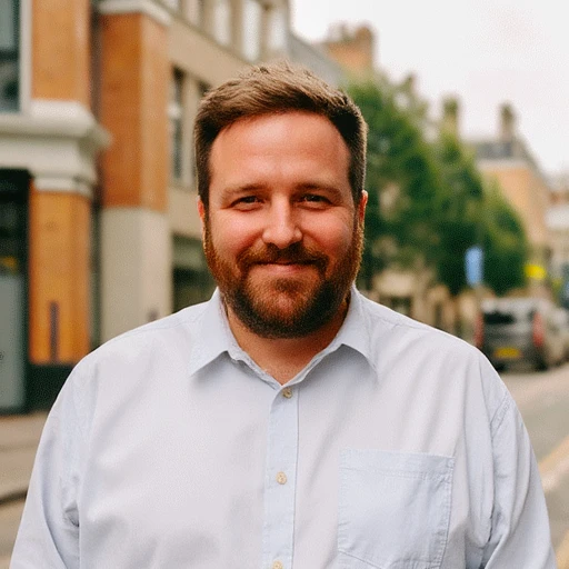 A photograph of the head and shoulders portrait of an average-looking male web developer in his thirties, standing on a street and wearing a button-up shirt, smiling at the camera. He has short brown hair with dark blonde sides and a full beard.